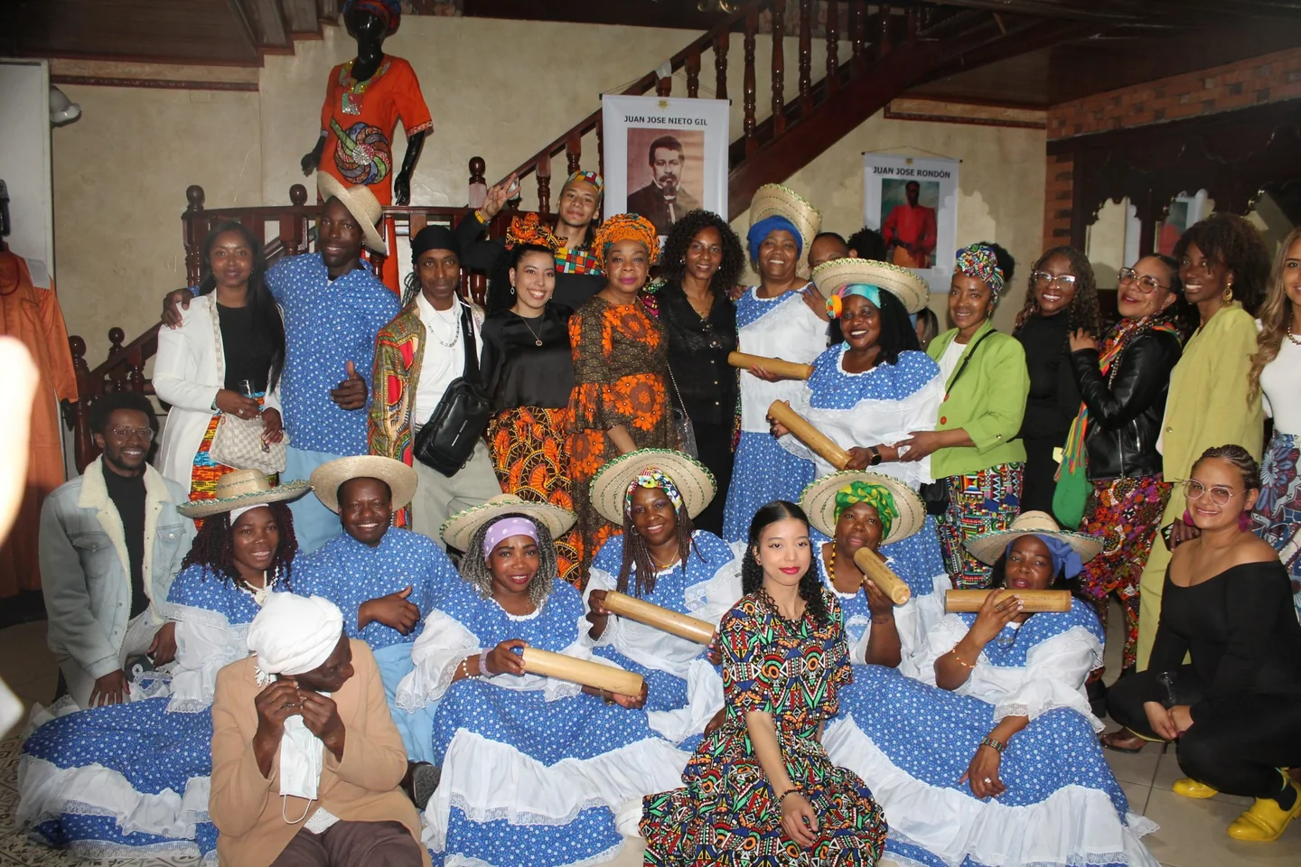Grupo de personas posando en un interior, varias con vestidos azules y blancos, sombreros e instrumentos de percusión.
